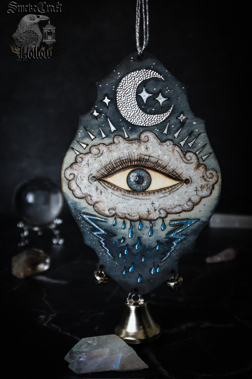 Art depicting an all seeing eye in a rain cloud with lightning bolts and a silver moon and blue colouring. Witch bell door hanger for protection spells and magick.