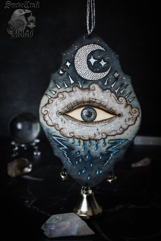 Art depicting an all seeing eye in a rain cloud with lightning bolts and a silver moon and blue colouring. Witch bell door hanger for protection spells and magick. 